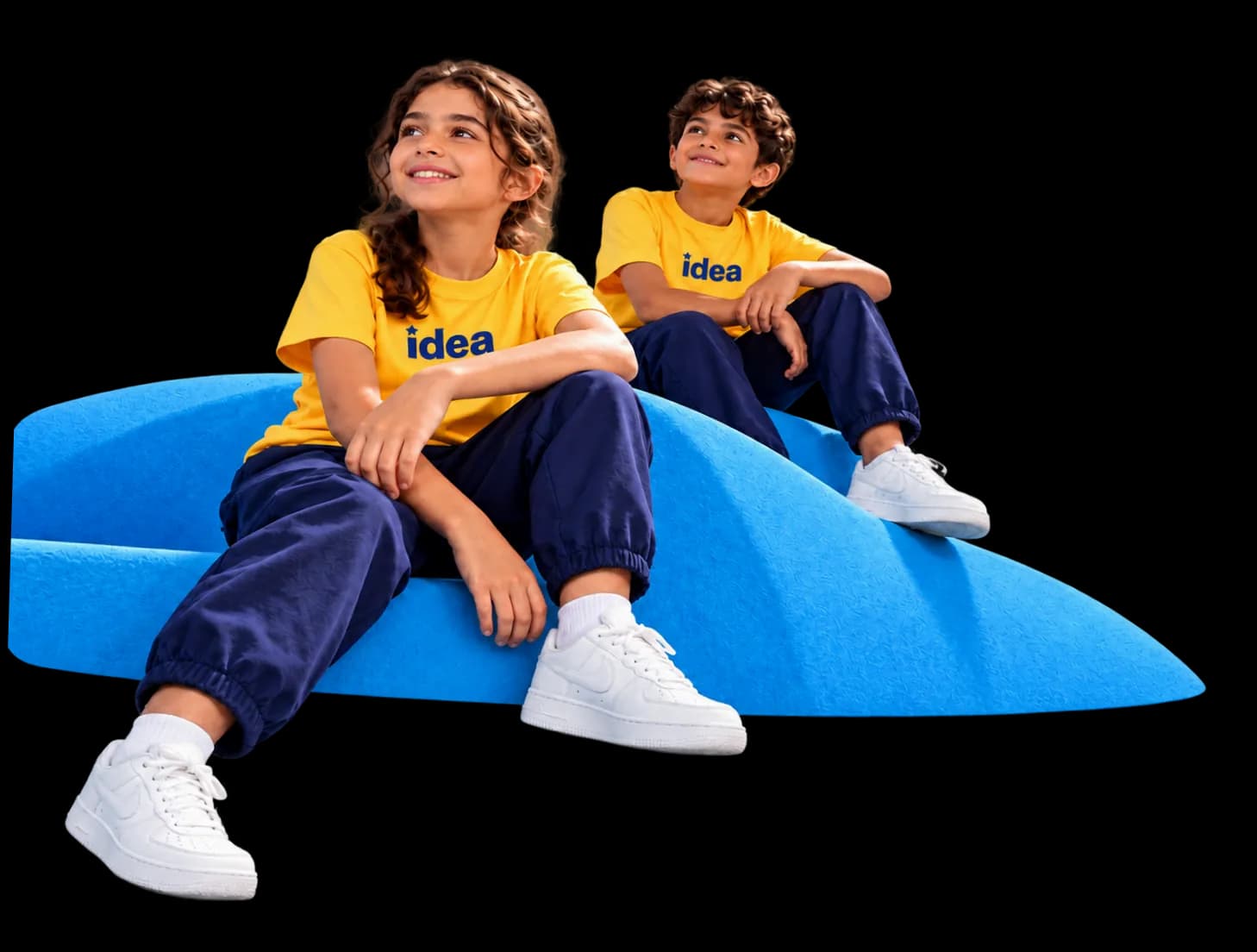طفلان مبتسمان بقميصين أصفرين عليهما شعار IDEA يجلسان على تلة زرقاء